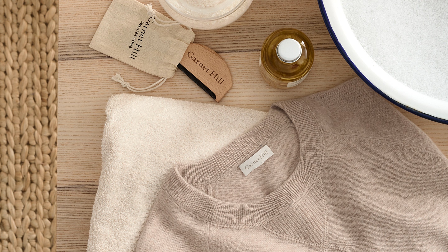 A tan cashmere sweater on a table with a bowl of soapy water and a sweater comb.