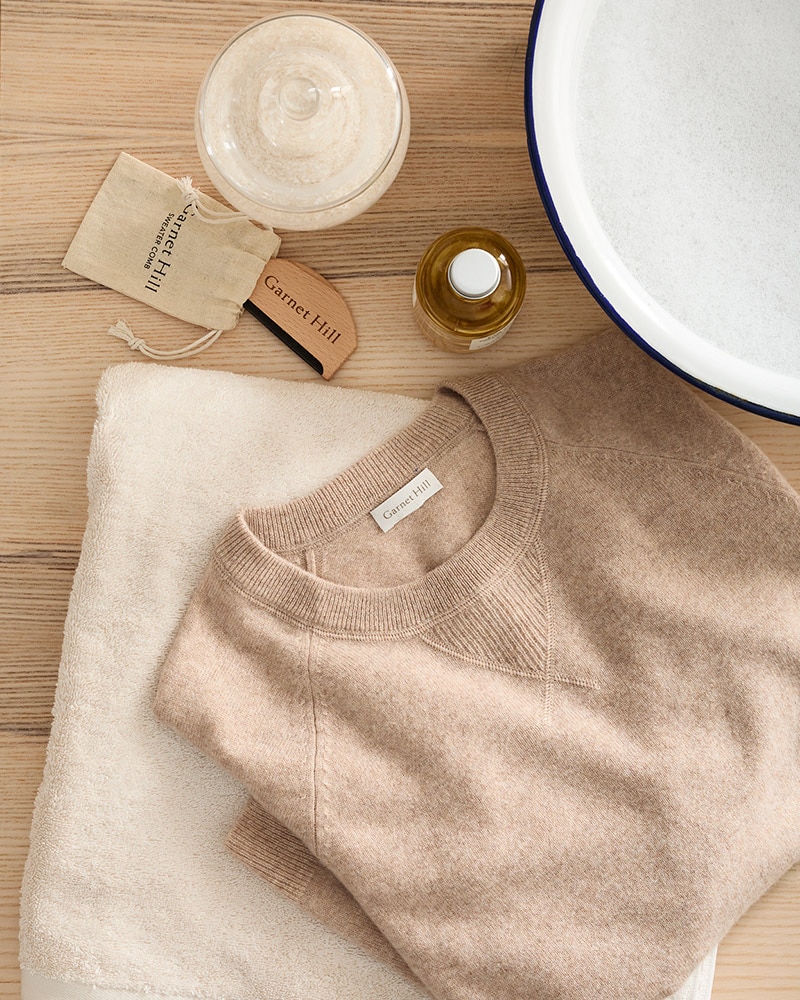 A tan cashmere sweater on a table with a bowl of soapy water. Learn how to care for cashmere.